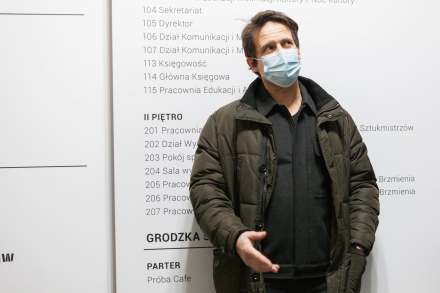 Mężczyzna stoi pod ścianą, na której rozpisany jest plan budynku przy ulicy Grodzkiej 7. Patrzy do góry, a prawą rękę ma wystawioną w stronę aparatu.