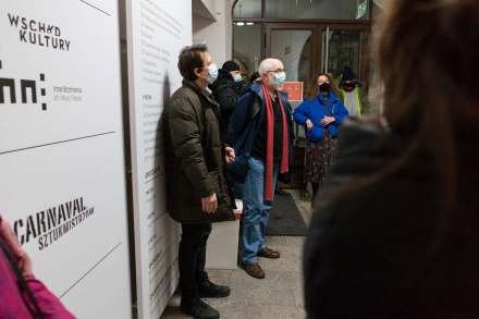 Ludzie stoją na korytarzu Warsztatów Kultury przy ulicy Grodzkiej 7 i słuchają wystąpienia otwierającego wystawę.