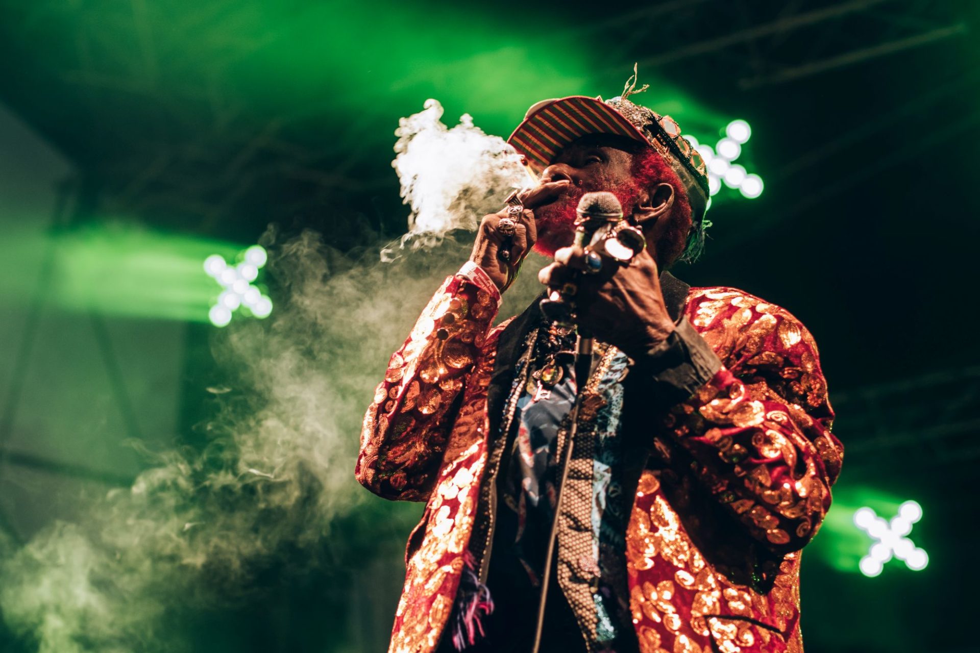 Na zdjęciu artysta reagge podczas koncertu. Afroamerykanin w podeszłym wieku ubrany w wielobarwny, oryginalny strój. Jego broda i włosy pofarbowane są na czerwono. Fotograf uchwycił moment, gdy mężczyzna pali papierosa. W tle Zielone światła sceniczne.