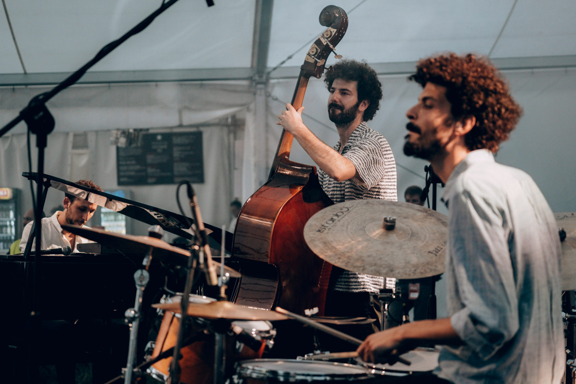 Na zdjęciu trzech muzyków o egzotycznej urodzie. Mężczyźni grają koncert w namiocie, widać takie intstrumenty jak perkusja, wiolonczela, fortepian.