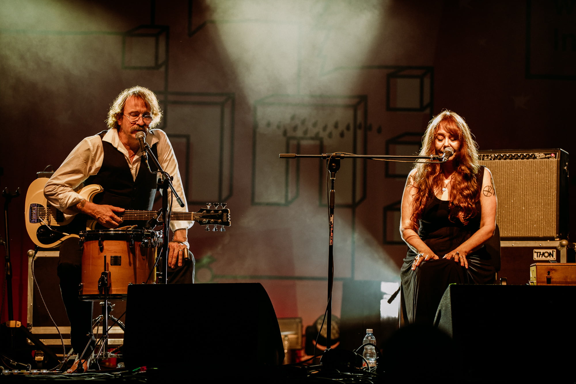 Obraz przedstawia dwoje muzyków, mężczyznę i kobietę, śpiewających do mikrofonu na scenie podczas koncertu.