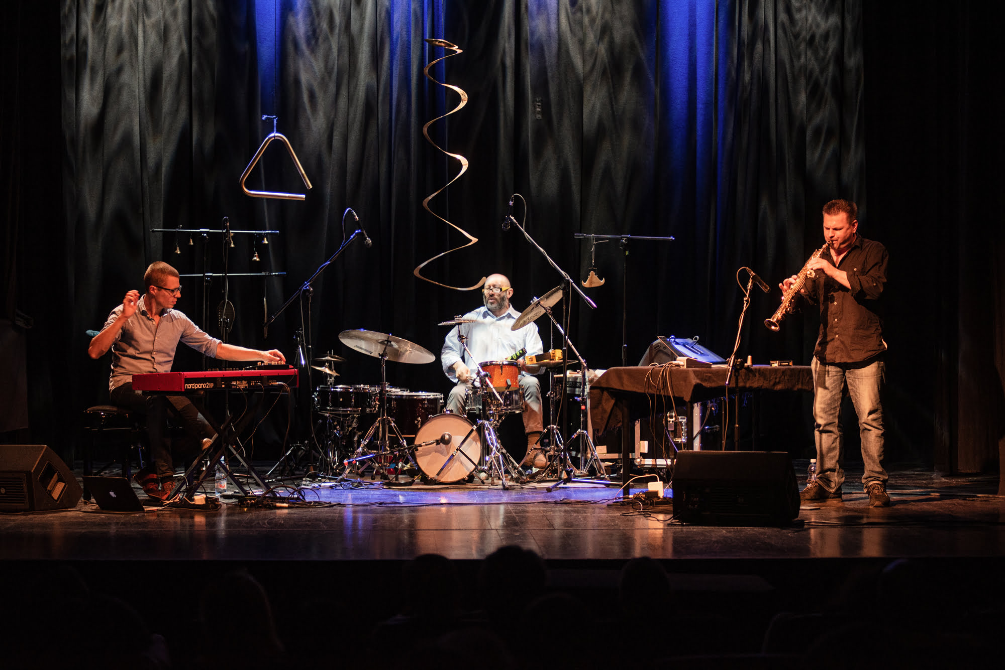 Zdjęcie kolorowe ze sceny, podczas koncertu. Przedstawia trzech muzyków:, od lewej strony siedzi pierwszy przy klawiszach, w środku