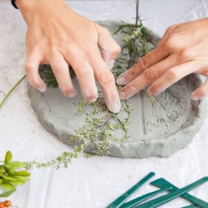 zdjęcie dłoni wykonującej prace plastyczne - odciskujących roślinę w glinie. obok odcisku leżą muszelki, szyszka i inne rośliny
