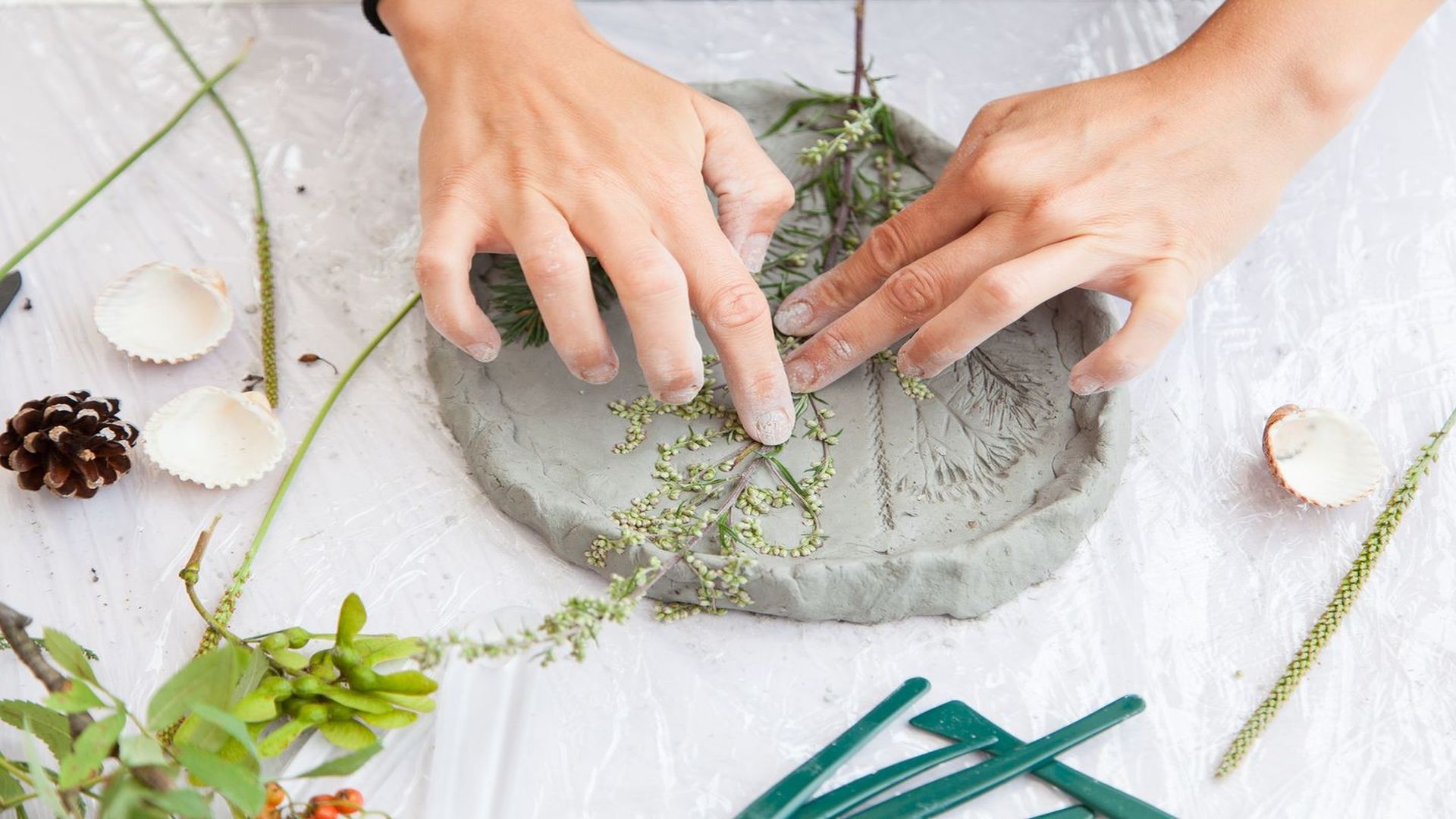 zdjęcie dłoni wykonującej prace plastyczne - odciskujących roślinę w glinie. obok odcisku leżą muszelki, szyszka i inne rośliny