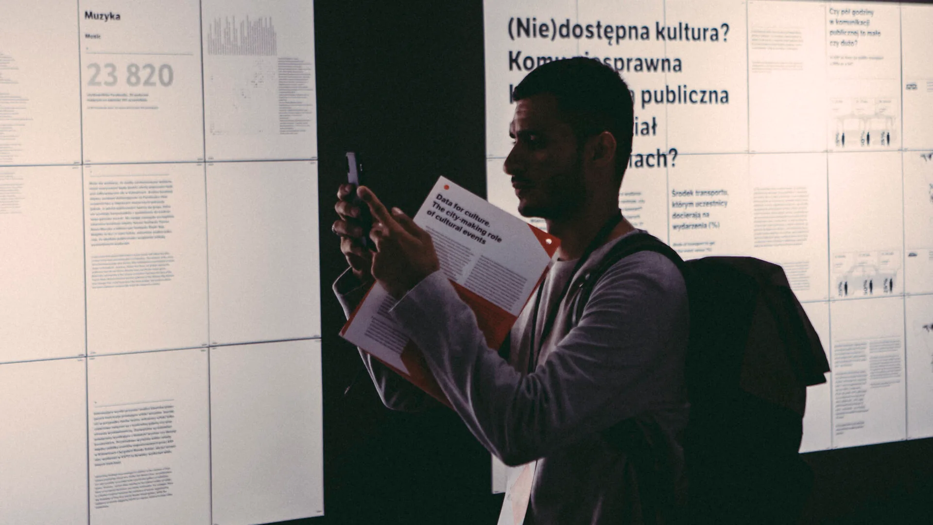 Zdjęcie pokazujące mężczyznę robiącego zdjęcie telefonem komórkowym. Znajduje się w instytucji kultury i ogląda ekspozycję. W ręku ma folder informacyjny.