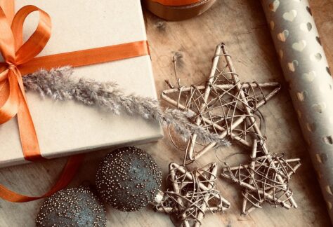 na drewnianym blacie leżą ozdoby choinkowe - dwie szklane bombki i 3 gwiazdki z gałązek, obok tekturowe pudełko z pomarańczową kokardką i rolka ozdobnego papieru. Wszystko w ciepłej, złocistej tonacji
