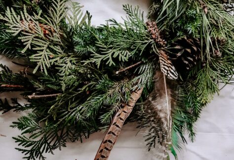 w kadrze widoczne pół wianka z gałązek iglastych, głównie żywotnika z powtykanymi szyszkami i ptasimi piórkami, wianek leży na jasnej płóciennej tkaninie