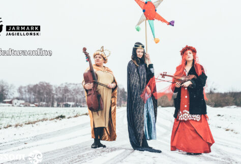 Trzy kobiety w egzotycznych, połyskujących strojach stoją na polnej drodze pokrytej śniegiem. Na głowach mają korony, a w rękach trzymają skrzypce, gwiazdę kolędniczą - atrybut kolędników i basy.. W tle domy i drzewa. W prawym górnym rogu logo Jarmarku Jagiellońskiego oraz hasztag kulturaonline. Autorką zdjęcia jest Marianna Łakoma, grafikę do promocji wydarzenia przygotowała Ilona Wałęcak