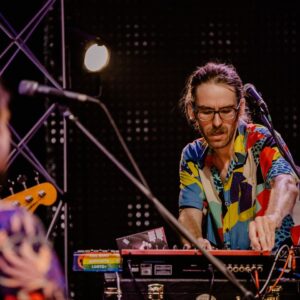 dwóch mężczyzn podczas koncertu. jeden, ubrany w kolorową koszulę pochyla się nad mikserem do muzyki, drugi stoi