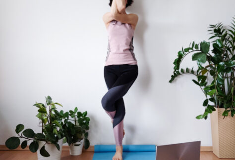 Na zdjęciu, w otoczeniu roślin doniczkowych, osoba w stroju gimnastycznym stoi na jednej nodze na niebieskiej macie, ma przed sobą laptopa. Unosi skrzyżowane ramiona zasłaniając twarz.