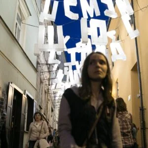 Zdjęcie wykonane podczas Nocy Kultury.Ludzie w wąskiej uliczce, nad ulicą zawieszone duże białe, święcące litery