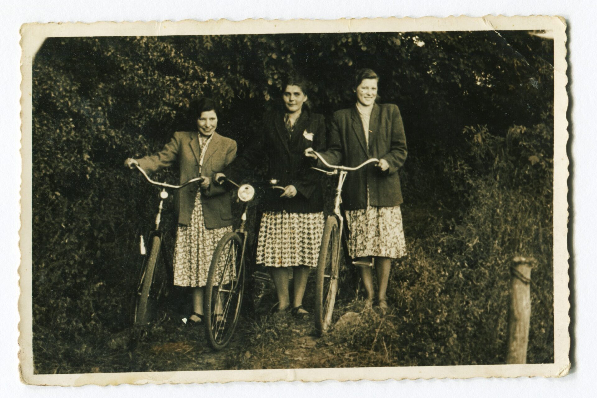 Zdjęcie archiwalne, sepia, widzimy trzy młode, uśmiechnięte dziewczyny ubrane w spódnice i marynarki. Każda z kobiet po swojej prawej stronie trzyma rower. Zdjęcie wykonane na tle drzew