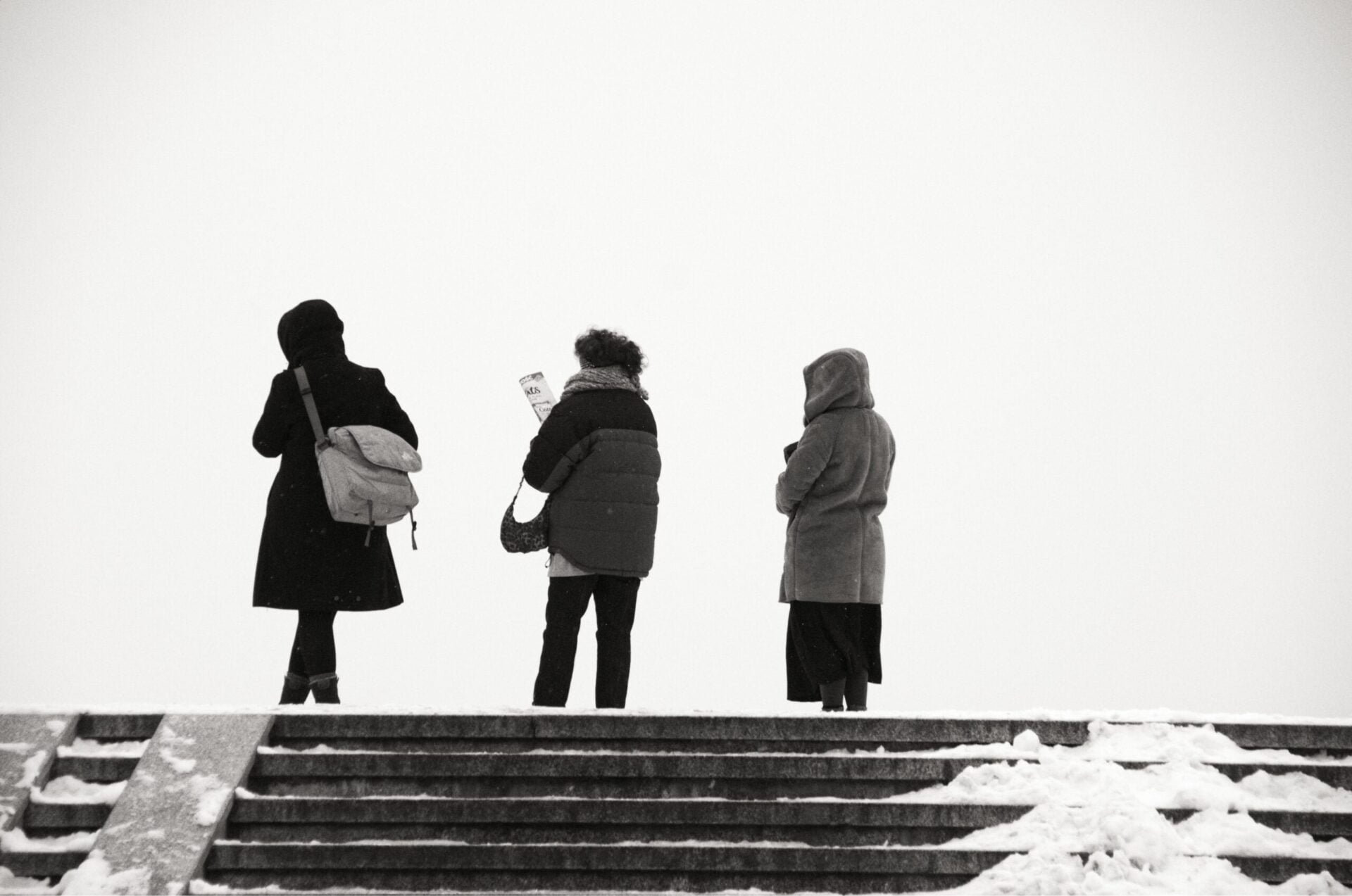 Zdjęcie czarno białe. Jest zima, śnieg, Na szczycie schodów stoją trzy osoby odwrócone tyłem do fotografii.