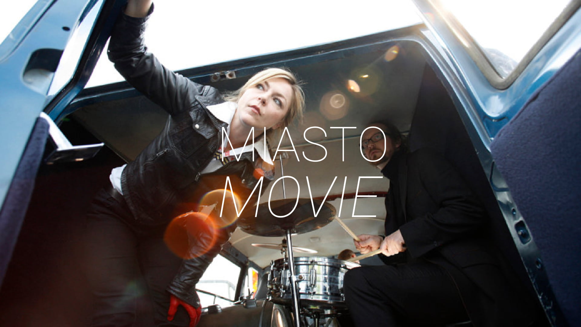 Zdjęcie kobiety i mężczyzny przy perkusji w bagażniku vana. Na środku napis Miasto Movie.