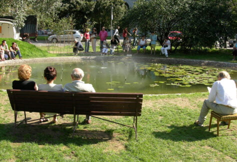Na zdjęciu widać mały staw wokół którego na ławkach lub na ziemi siedzą ludzie