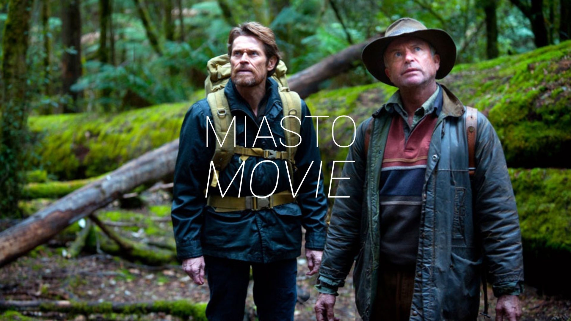 Dwaj mężczyźni idą przez dżunglę. Mają plecaki, za nimi jest powalone drzewo. Na środku logo Miasta Movie.