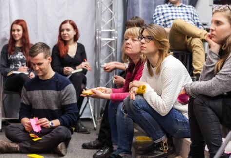 A group of students sitting on chairs and on the floor. Some are taking notes.