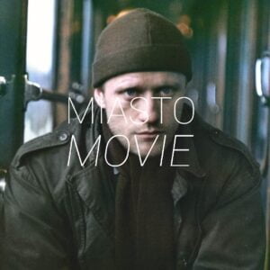 Kadr z filmu. Mężczyzna w kurtce zimowej, czapce i szaliku siedzi na środku wagonu pociągu i patrzy przed siebie. Na środku logo Miasta Movie.