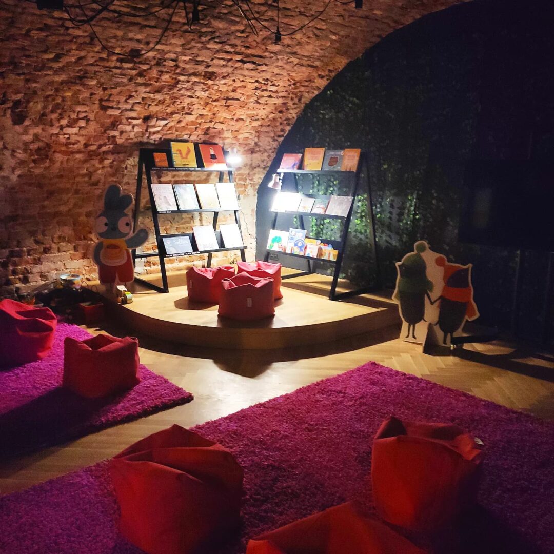 274576825_292532006315651_449277020659883120_n Wnętrze Cafe Próba, która obecnie służy za świetlicę dla dzieci z Ukrainy. Na różowych dywanach czerwone pufy do siedzenia. W tle dwa regały z książkami dla dzieci w języku ukraińskim.