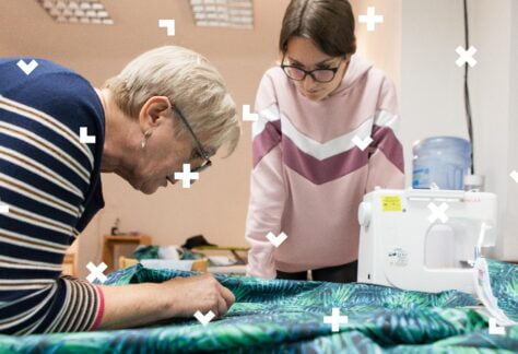 Na zdjęciu starsza kobieta pochyla się nad ciemnozielonym materiałem z motywem liści. Jej czynnościom przygląda się młoda dziewczyna w różowej bluzie. Drobne elementy graficzne przypominające bloki z gry tetris porozrzucane są na całym obrazku.