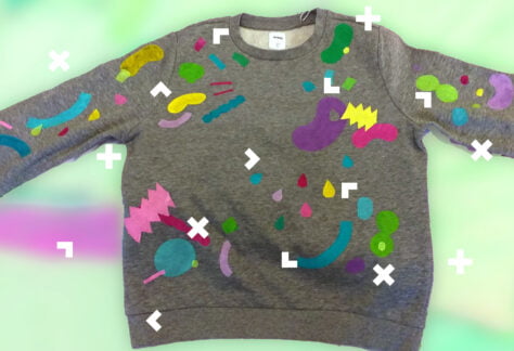 Na zielonym tle szara bluzka dziecięca pomalowana we wzory geometryczne różnymi kolorami.