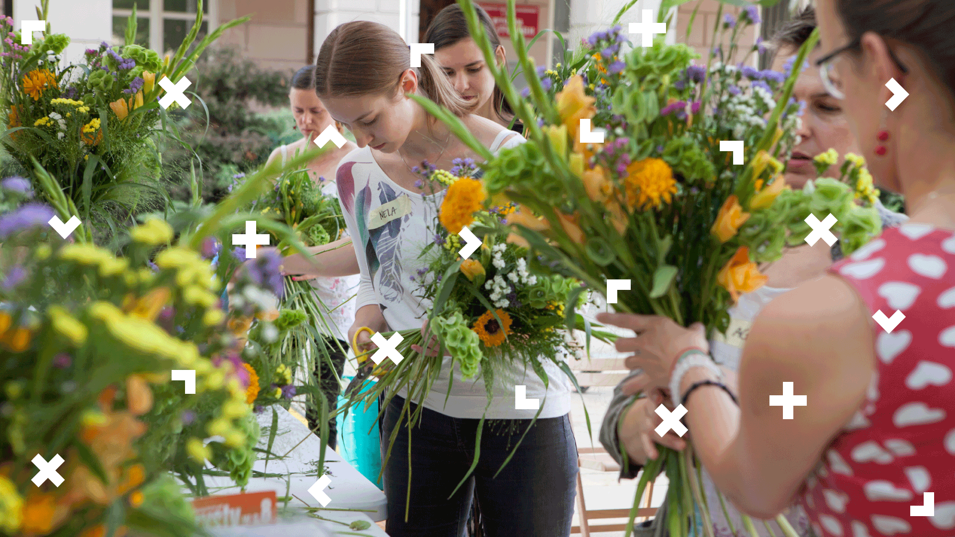 Grupa kobiet stoi przy stole pełnym kwiatów. Część z nich trzyma w ręce gotowe bukiety.