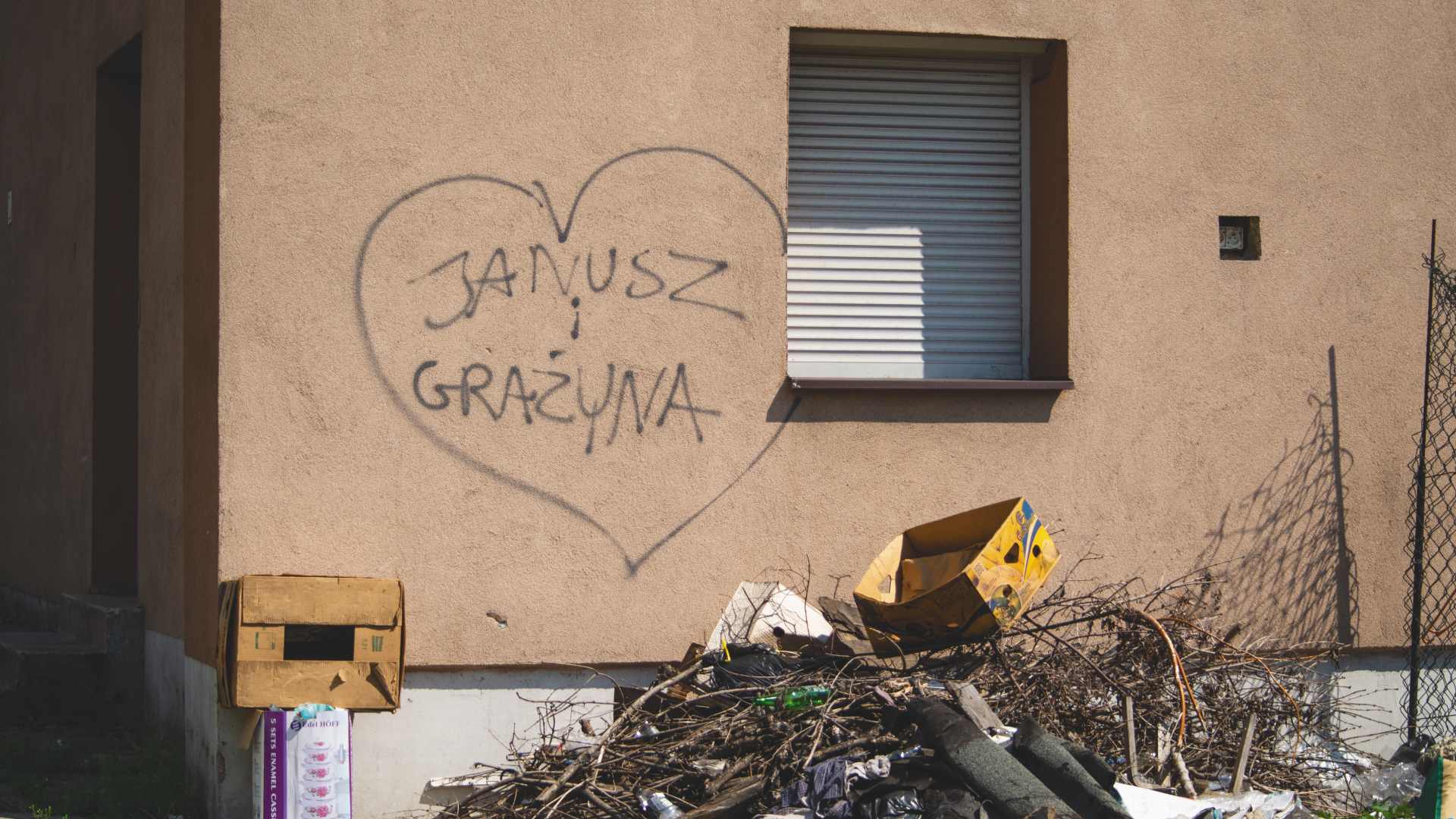 Na elewacji bloku mieszkalnego znajduje się serce z napisem: Janusz i Grażyna. Pod oknem leży gruz o śmieci.