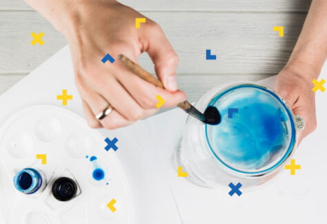 Kobieta trzyma w dłoni pędzelek, który moczy w wodzie. Na stole stoją paletka i farby.