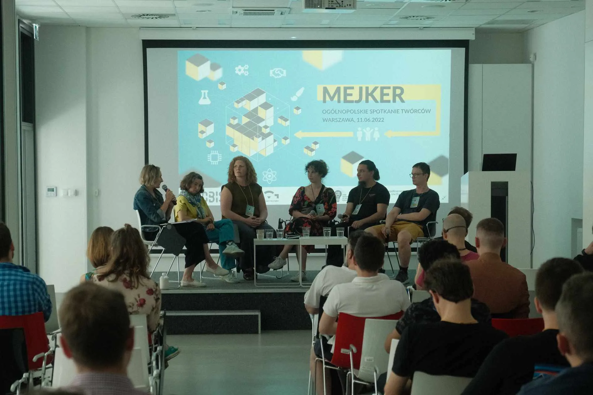 Sześć osób siedzi wokół stołu na podium. Za nimi wyświetlana jest prezentacja ze slajdem z napisem "Mejker". Widownia przysłuchuje się dyskusji.