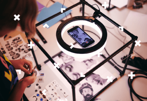 Na szklanej skrzyni leży telefon z włączonym aparatem skierowanym na zdjęcia znajdujące się w skrzyni. Kobieta stoi obok, w ręce trzyma monety.