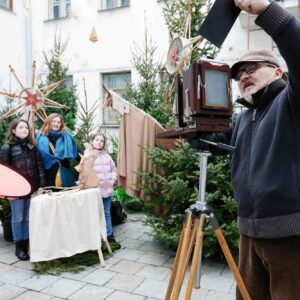 Roman Krawczenko stoi na patio Warsztatów Kultury z aparatem analogowym. Dookoła są choinki świąteczne, a na środku stoją kobiety gotowe do zdjęcia.
