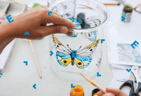 Stół z materiałami plastycznymi. Na stole sto duży słoik, na którym ktoś maluje farbami motyla.