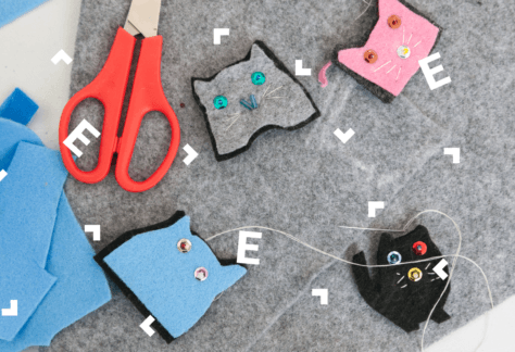 Na blacie leżą kawałki szarego i niebieskiego filcu, nożyczki oraz kotki wycięte z filcu, z wąsami z nitek i oczami z cekinów.