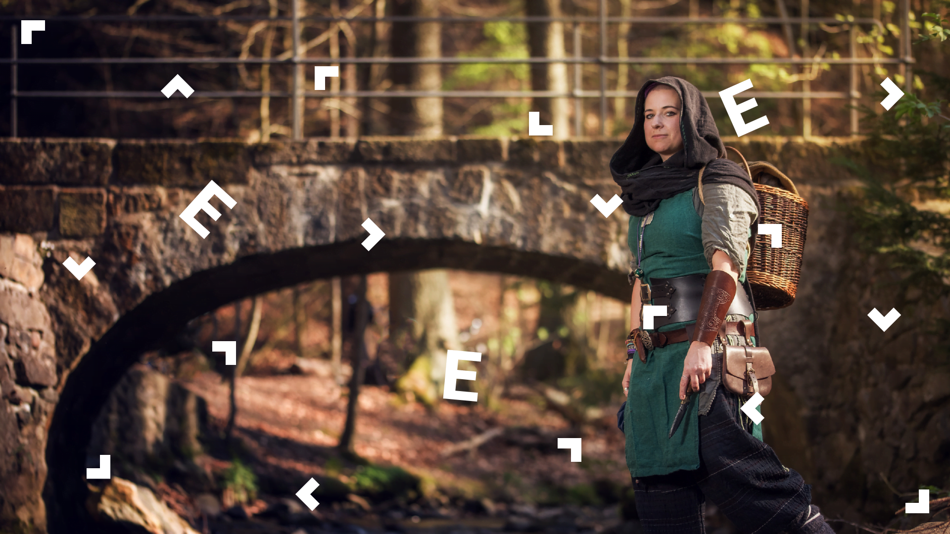 Kobieta przebrana w średniowieczny, zielony strój z kapturem stoi w lesie przy kamiennym moście.
