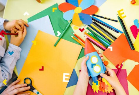 Trójka dzieci siedzi przy stole i sklejają kosmiczną rakietę. Na blacie leżą kartki kolorowego papieru, kredki i nożyczki.