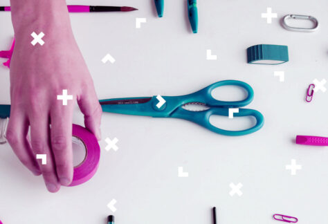 Na stole leżą nożyczki, spinacze i ołówek. Ktoś kładzie obok różową taśmę.