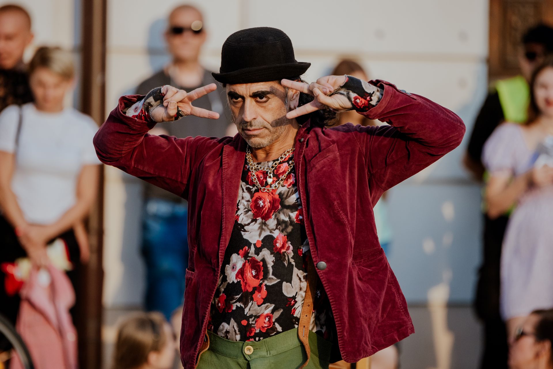 fot. Katarzyna Anna Kubiak (14) Artysta uliczny podczas występu. Mężczyzna jest ubrany w czerwoną marynarkę, czarny kapelusz i unosi ręce na wysokość swojej twarzy. Przyglądają mu się ludzie.