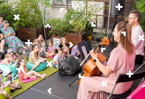 Dzieci siedzą na dywanie na patio Warsztatów Kultury. Mężczyzna i kobieta grają dla nich koncert.