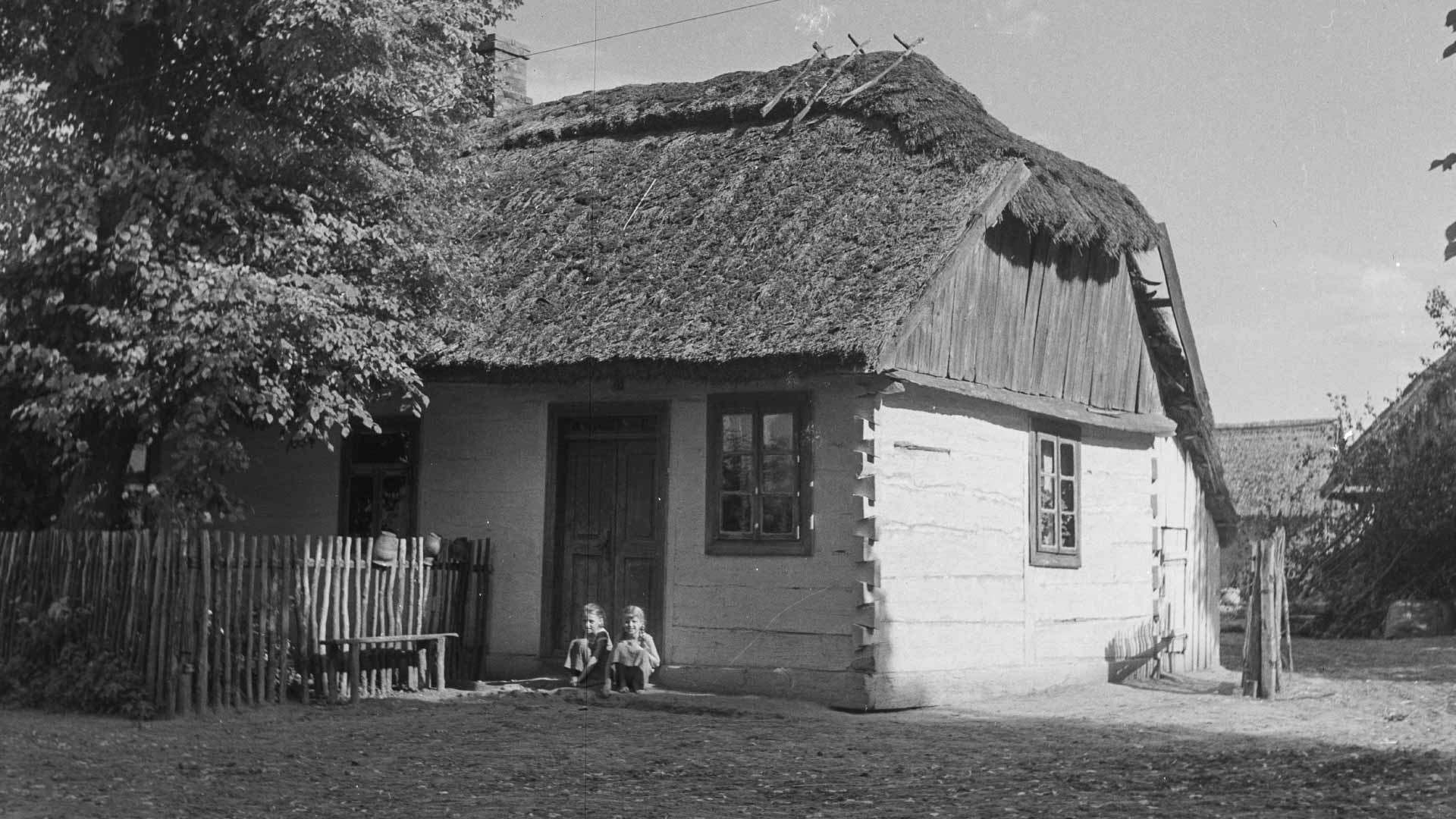 Czarno-biała fotografia sprzed lat. Przedstawia drewnianą chatę krytą strzechą. Pod drzwiami siedzi dwoje dzieci.