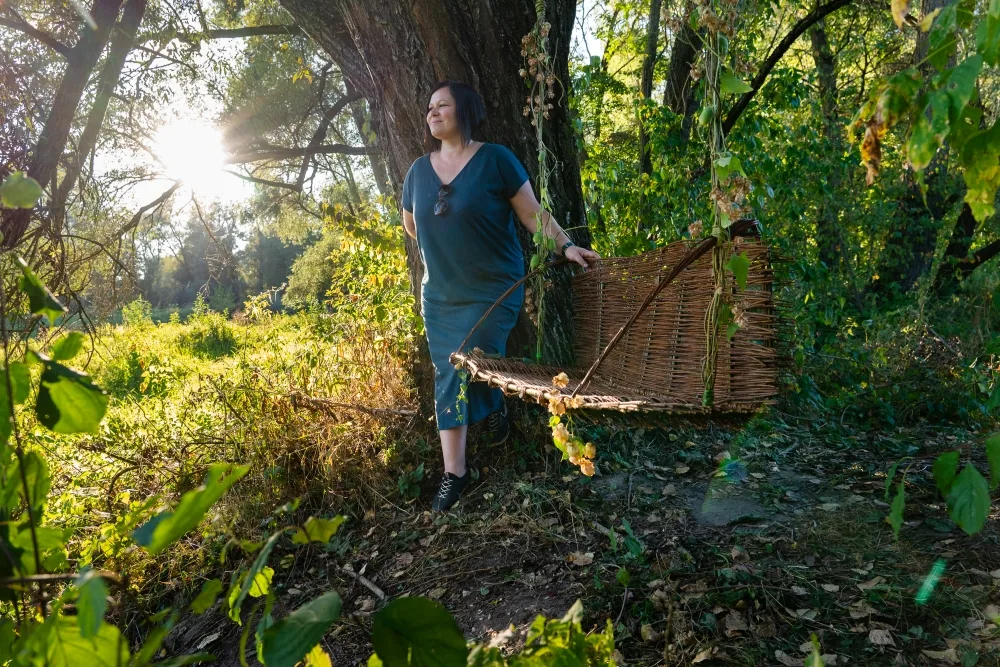 Kobieta stoi oparta o drzewo. Po jej prawej stronie znajduje się huśtawka upleciona z wikliny.