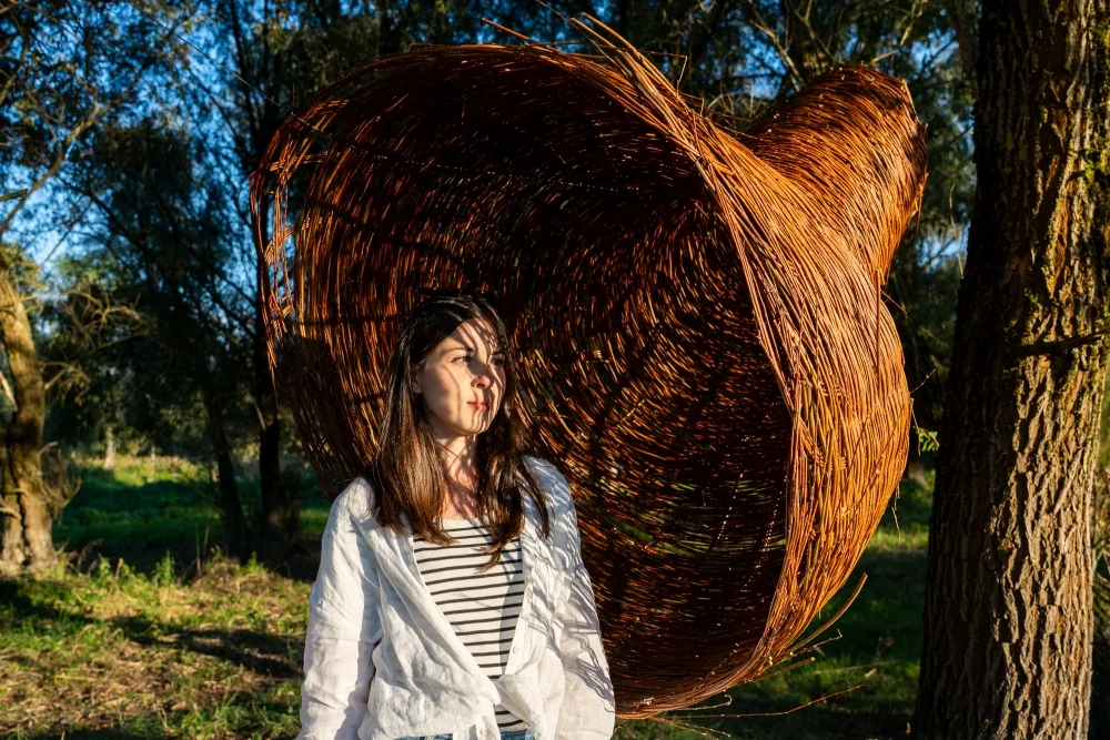 Na drzewie jest zawieszona tuba z wikliny. Przed nią stoi młoda dziewczyna.