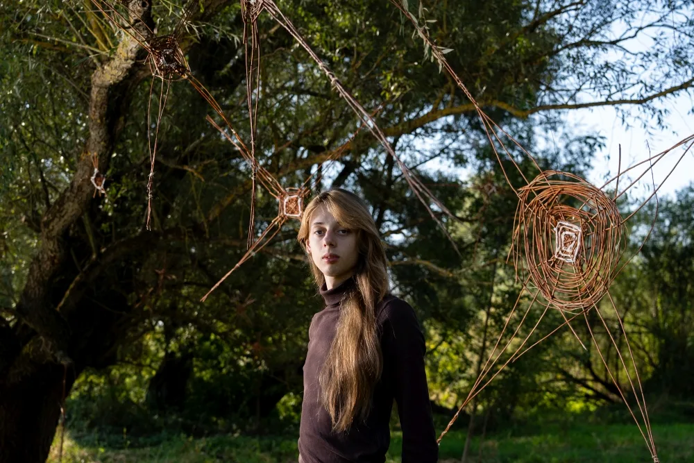 Dziewczyna stoi koło swojej pracy. To pajęczyna z wikliny zwisająca z drzewa.