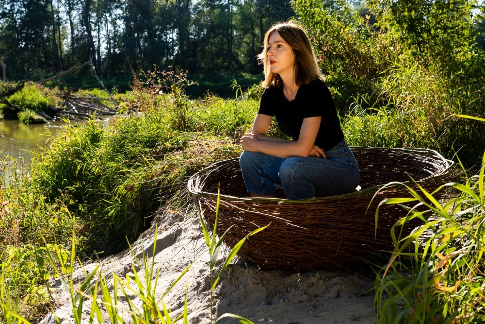 Kobieta siedzi w wiklinowej łódce. Dookoła są zarośla.