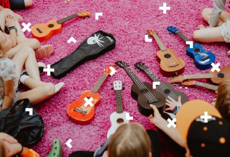 Dzieci siedzą na różowym dywanie. Przed nimi leżą ukulele.