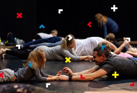 Dzieci i rodzice leżą na podłodze na brzuchu i trzymają się za ręce. Patrzą sobie w oczy.