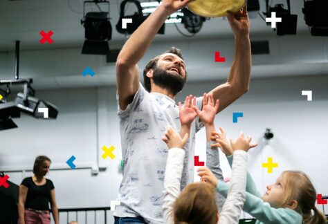 Mężczyzna trzyma nad głową bębenek, a dzieci skaczą dookoła niego.