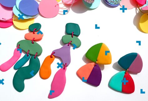 Na zdjęciu widoczne są dwie pary kolczyków zrobionych z kolorowych elementów o różnorodnych kształtach, połączonych za pomocą metalowych, różowych i złotych kółeczek. Nad kolczykami widać kolorową girlandę z okrągłych i kolorowych elementów, połączoną błyszczącą tasiemką.