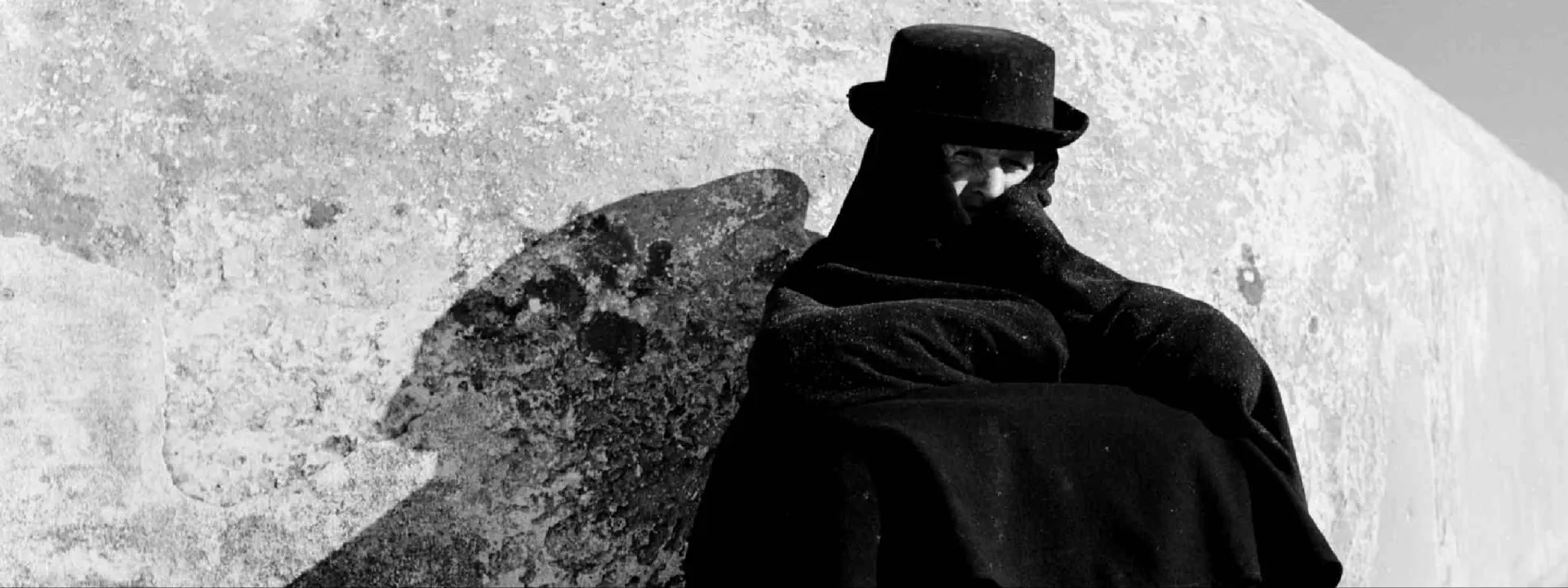 Czarno-białe zdjęcie osoby zawiniętej w koc i z kapeluszem na głowie. Widać tylko jego oczy. To prawdopodobnie mężczyzna.