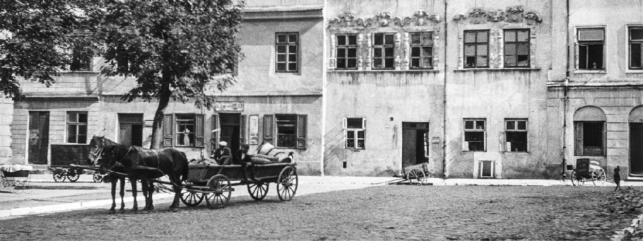 Czarno-biała fotografia. Dwaj mężczyźni siedzą na wozie zaprzężonym w dwa konie.