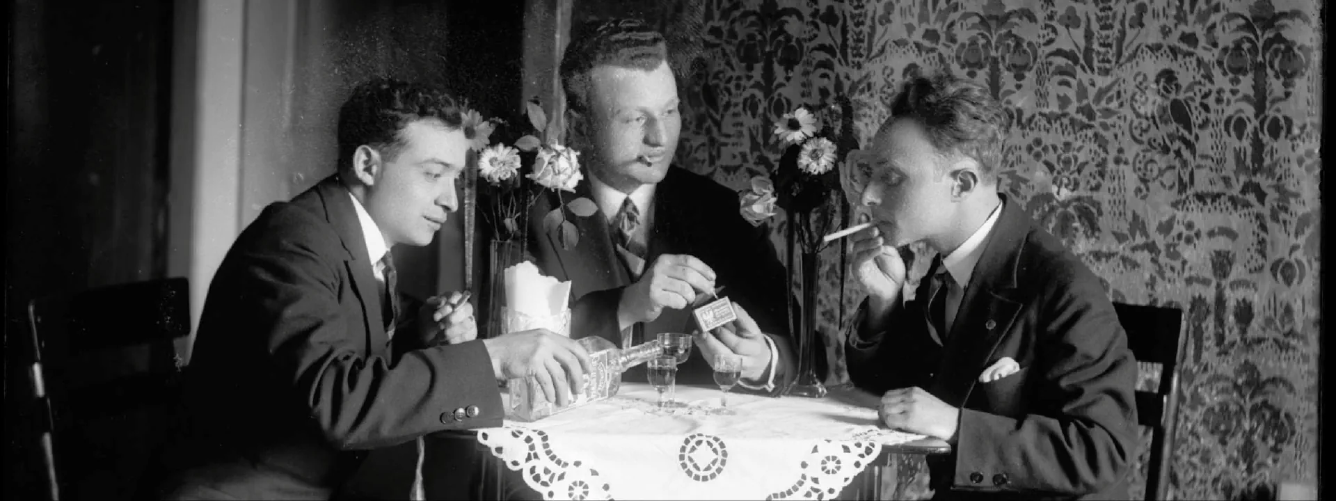 Czarno-białe zdjęcie trzech mężczyzn, którzy siedzą przy stoliku, piją alkohol i palą.